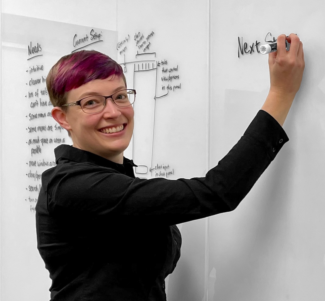 Sara, a white woman with short, magenta-colored hair, smiles at the camera while writing on a whiteboard