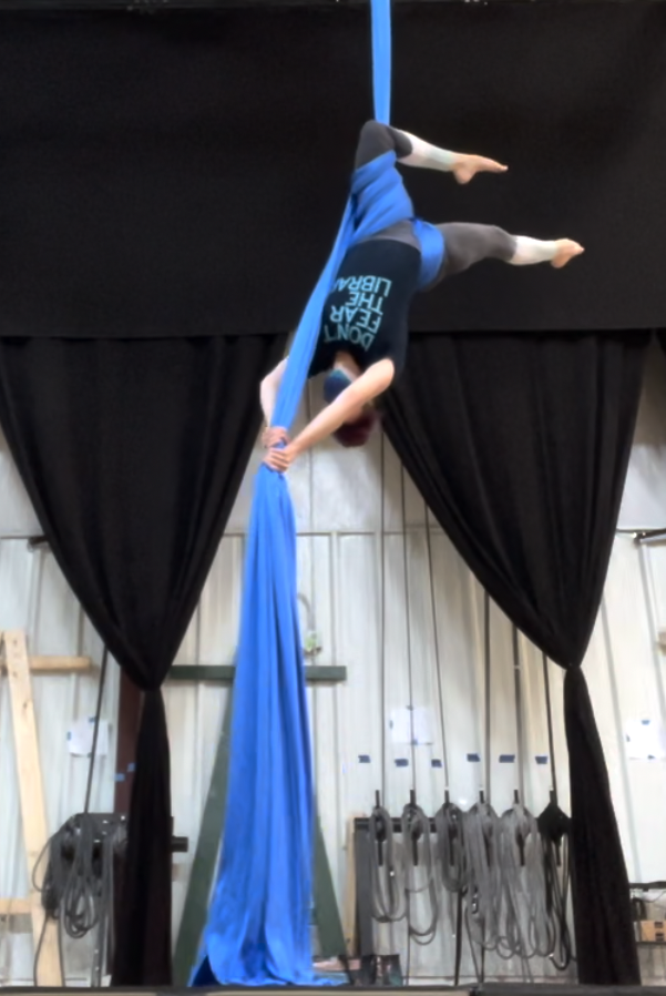 Photo of Sara on a blue fabric hanging from the ceiling. She is upside down; she has one leg wrapped with the knee hooked over the taut fabric and is holding the loose tail in both hands.