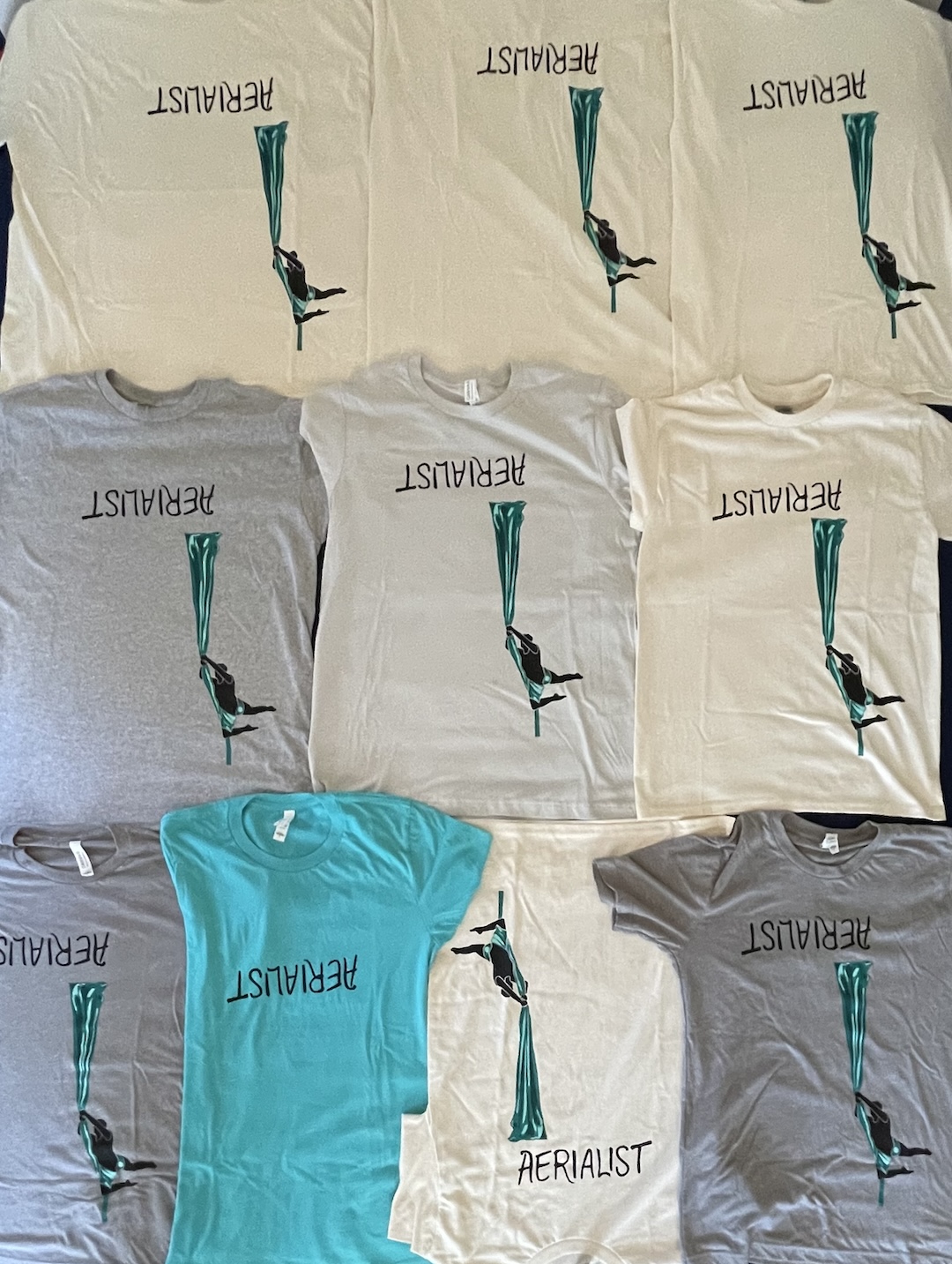 ten shirts laid flat, mostly grey or beige with one bright turquoise, printed with "aerialist" print (turquoise shirt only has the text, no image)